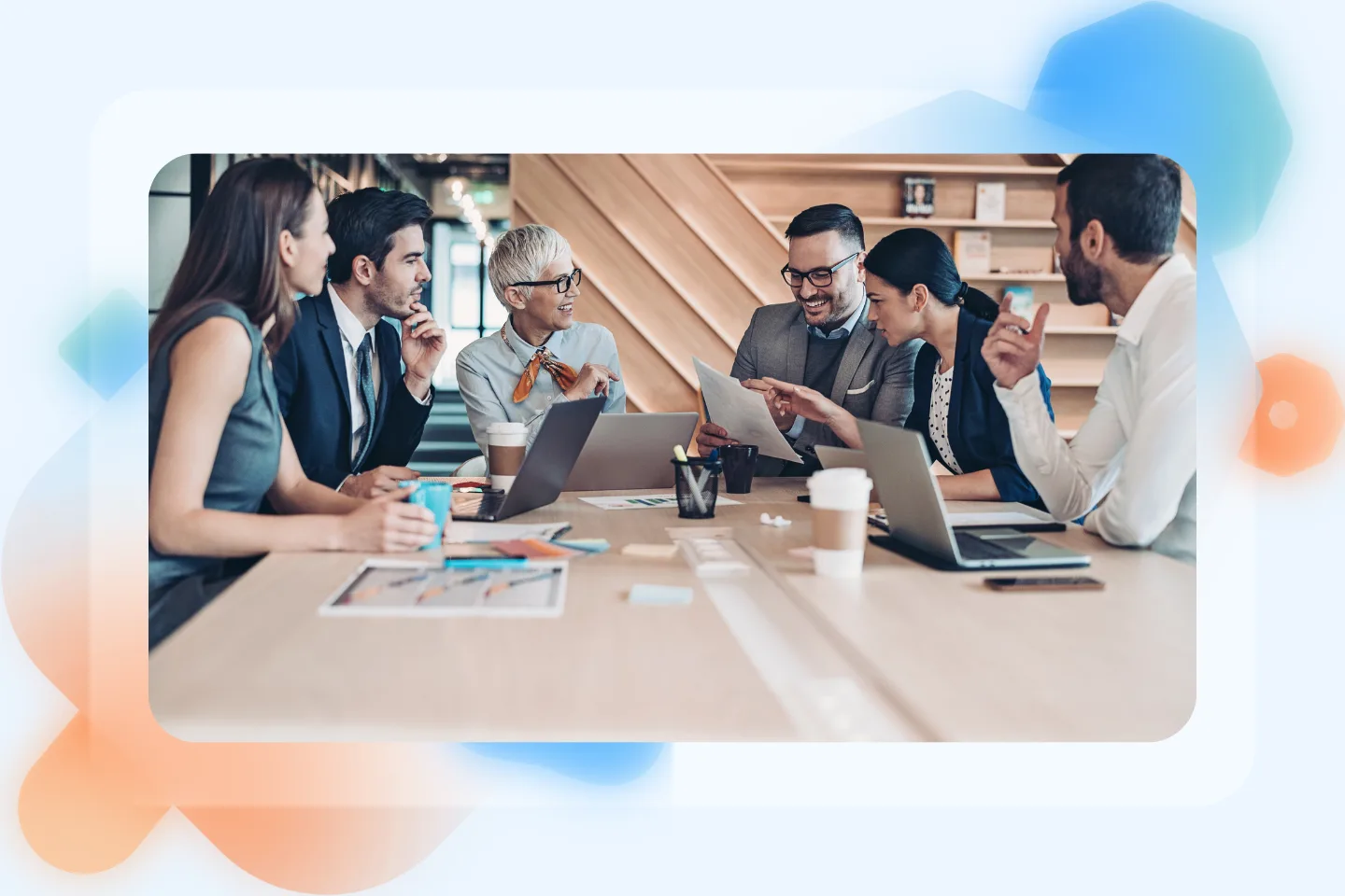 Six professionals in a meeting around a wooden table with laptops and coffee, reviewing documents already on the table.